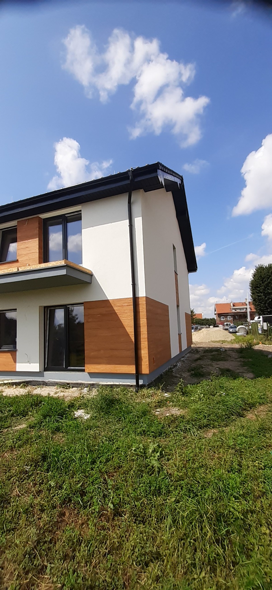 Nowoczesny dom jednorodzinny z białymi ścianami i drewnopodobną elewacją, czarne rynny, trawnik, błękitne niebo z chmurami w tle. Ujęcie z dołu, podkreślające bryłę budynku.