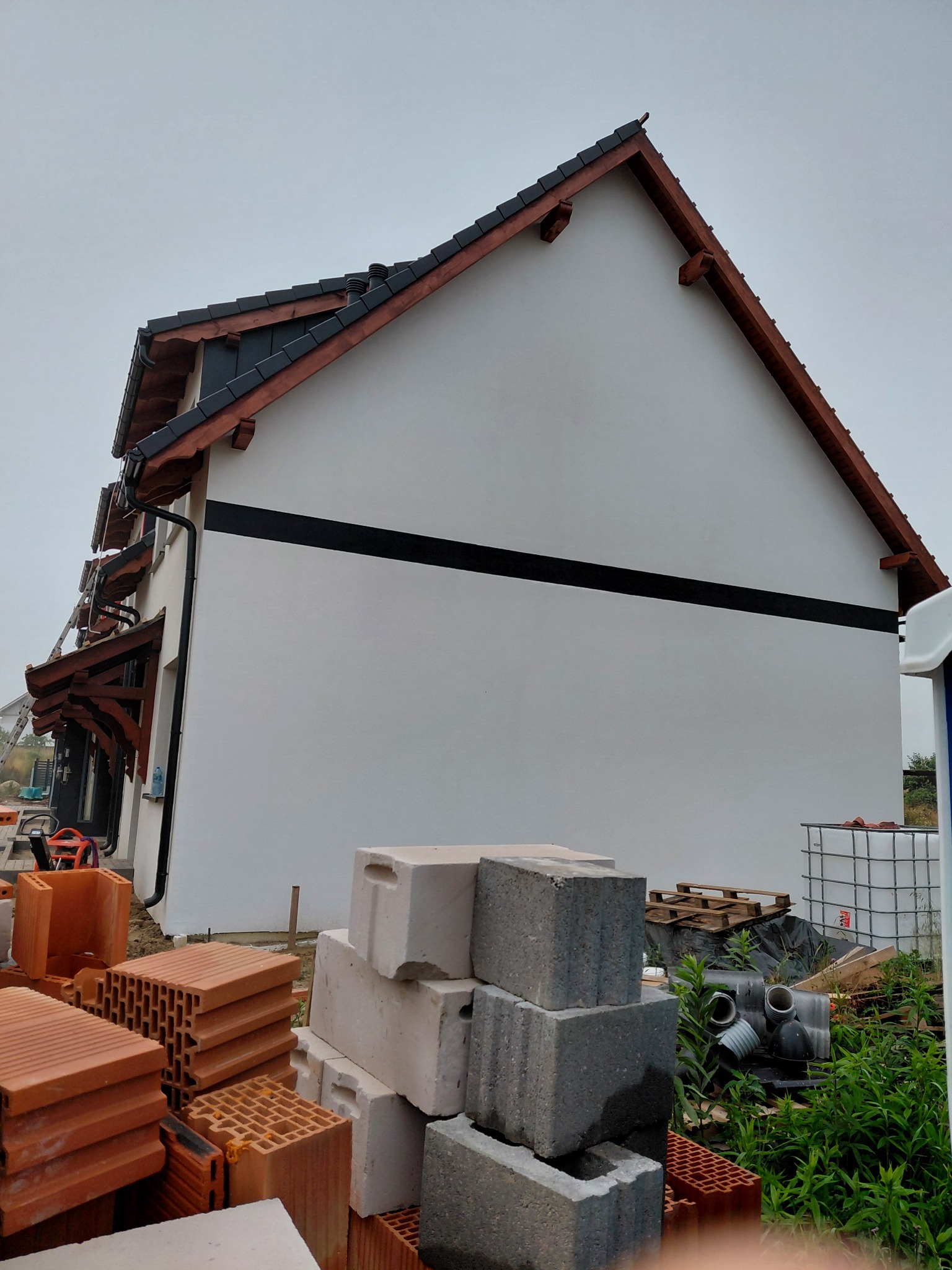 Ściana domu z wykończoną elewacją, widoczne cegły i materiały budowlane. Świeżo otynkowana ściana z czarnym pasem, brązowe elementy dachu.