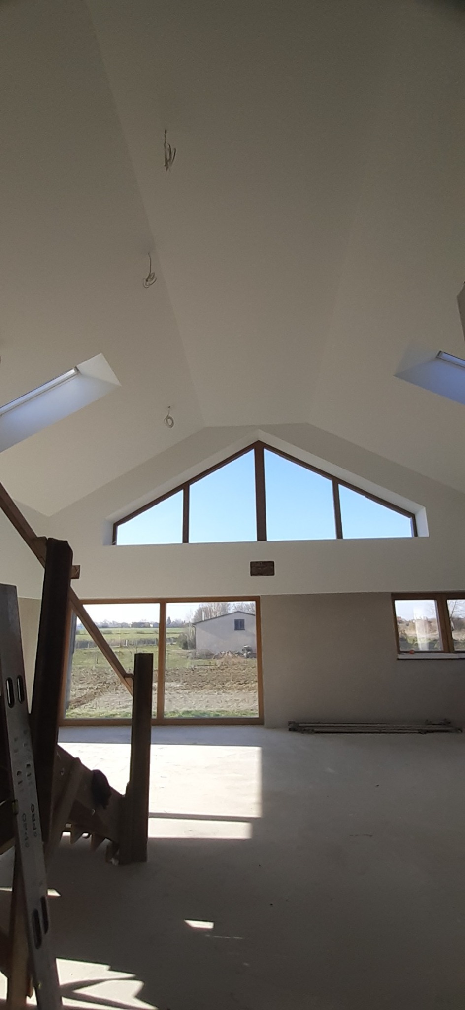 Wnętrze poddasza po remoncie: białe ściany i sufit, okna dachowe i panoramiczne z widokiem na zewnątrz. Widoczne przewody elektryczne, drabina i surowa podłoga.