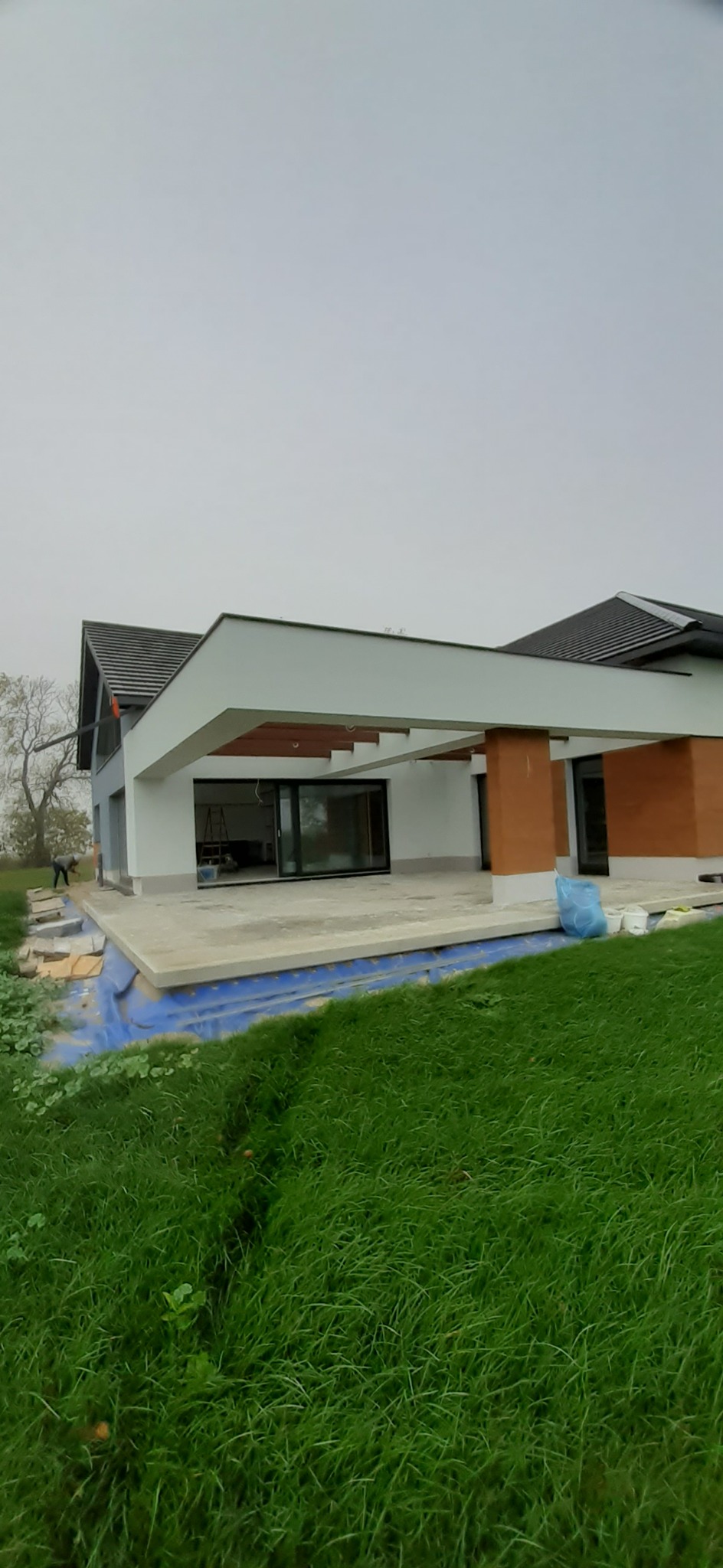 Nowoczesny dom jednorodzinny z płaskim dachem i dużymi przeszkleniami, taras w trakcie wykończenia, widoczne elementy izolacji fundamentów.