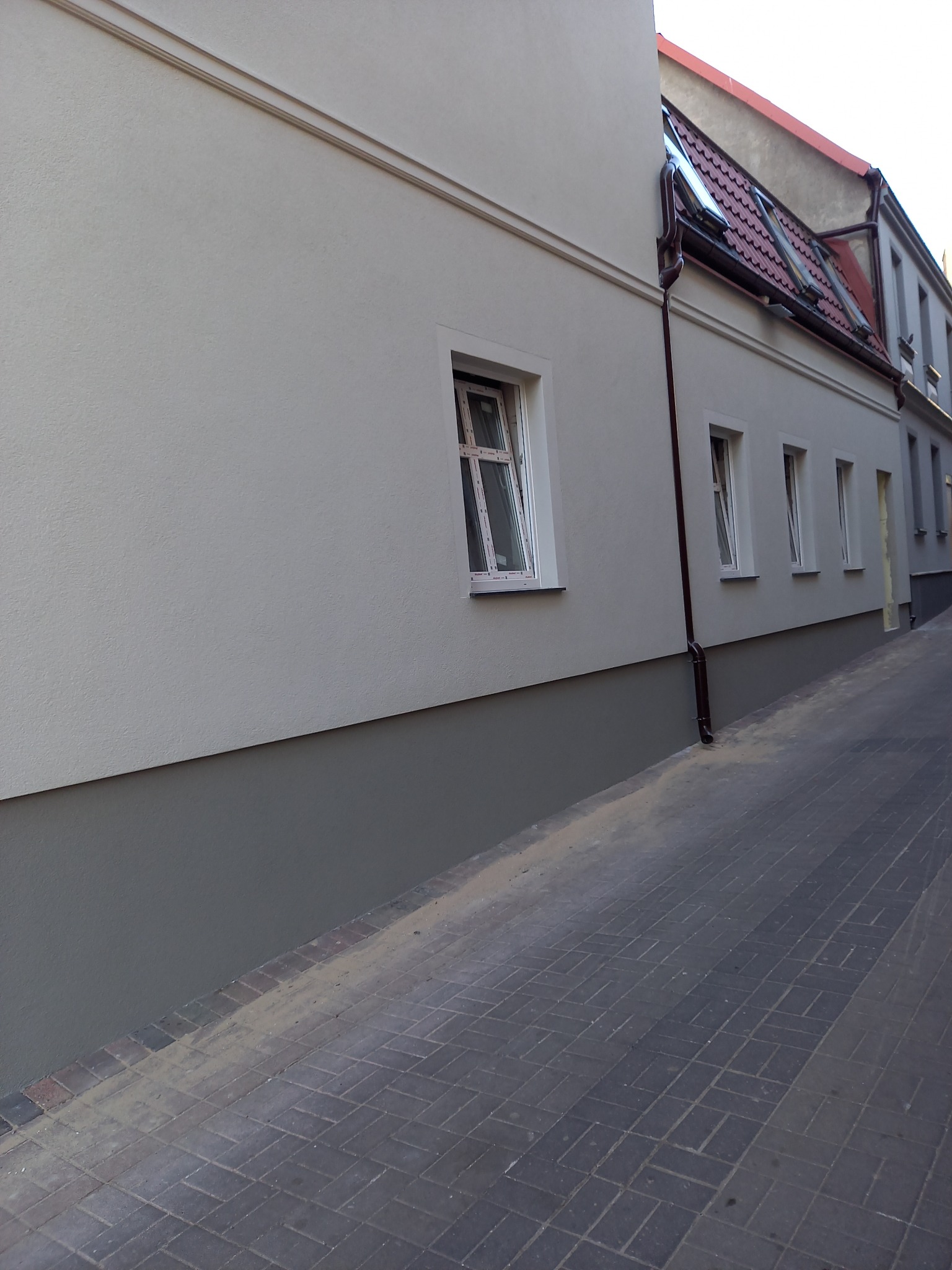 Ocieplony budynek z elewacją w jasnych odcieniach szarości, z widocznymi oknami i rynną. Dolna część budynku pomalowana na ciemniejszy odcień.