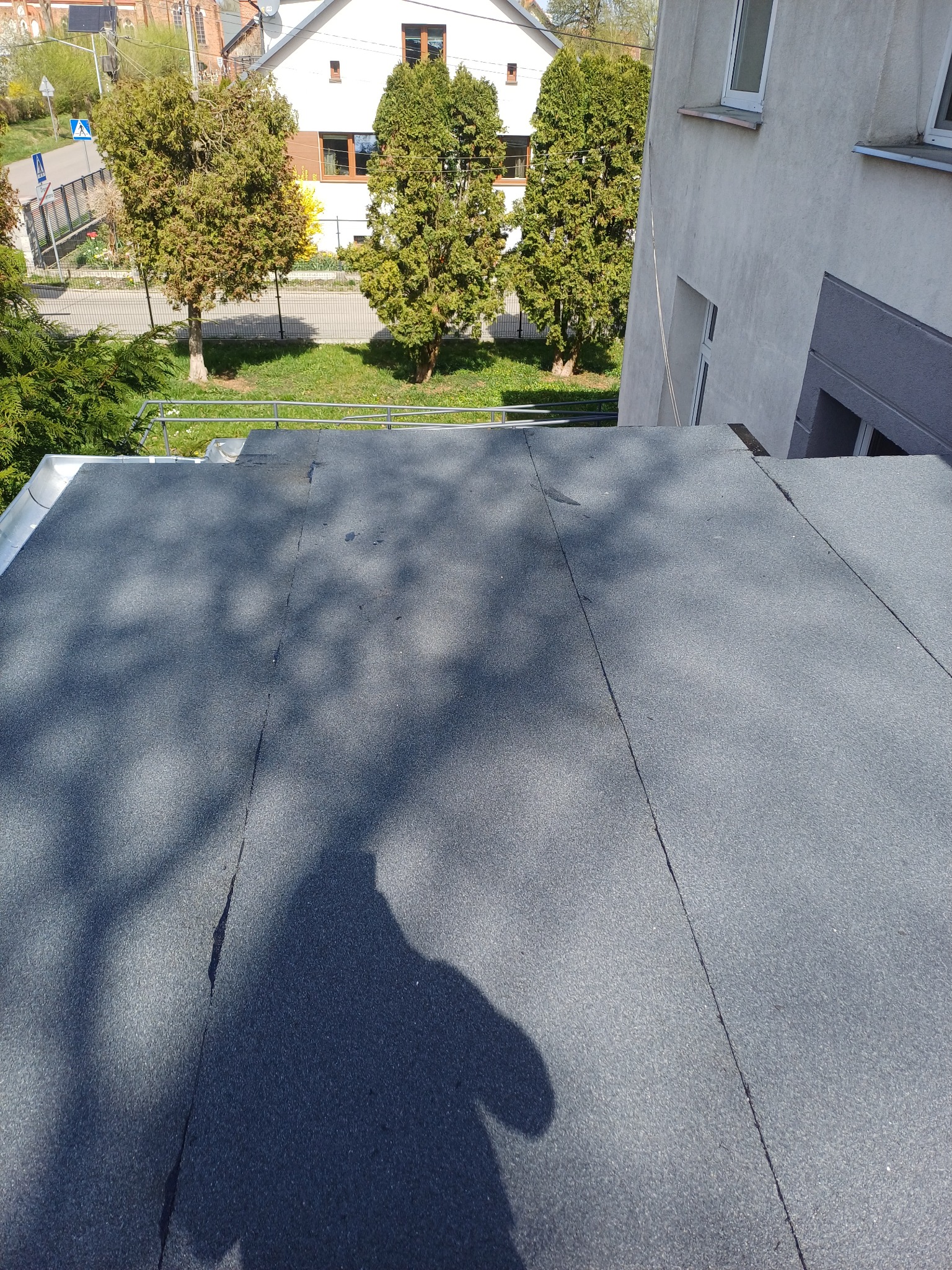 Szary, płaski dach pokryty papą na tle zieleni i budynków. Widoczny cień drzewa na powierzchni dachu. Kąt ujęcia z góry, podkreślający fakturę pokrycia.