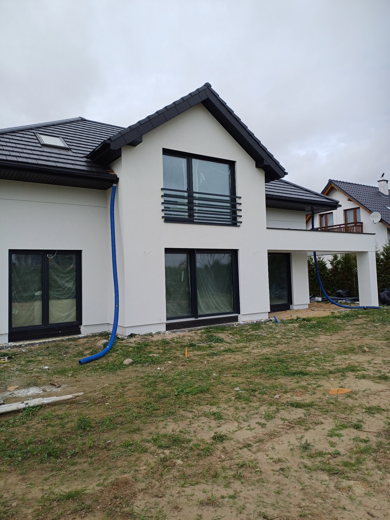 Nowoczesny dom z białymi ścianami i czarnym dachem, widoczne niebieskie węże do izolacji. Okna zabezpieczone folią, teren wokół domu przygotowany do zagospodarowania.
