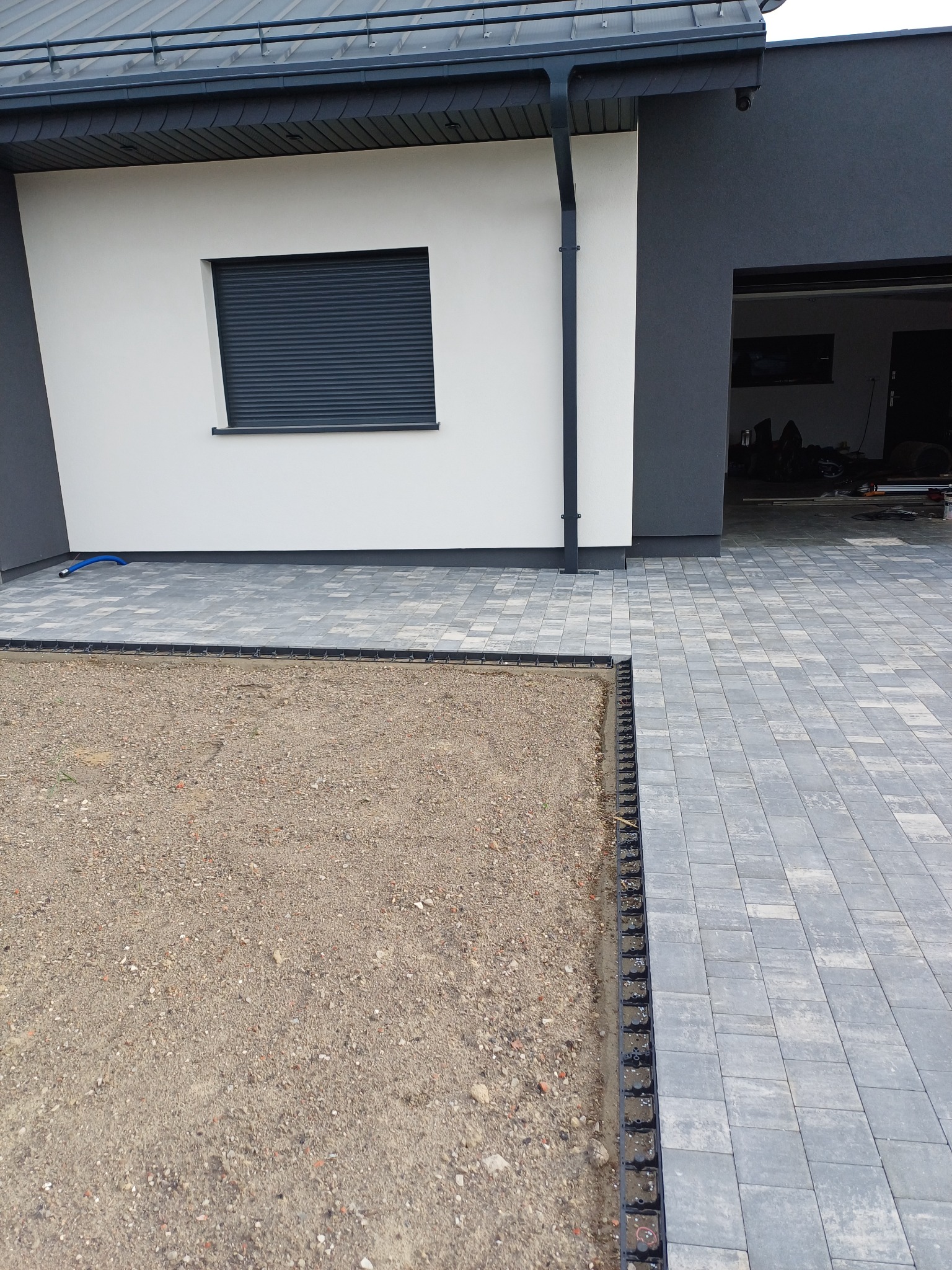 Nowoczesny dom z szarą elewacją i roletami zewnętrznymi. Wybrukowany podjazd przechodzi w teren przygotowany pod trawnik z obrzeżami. Widoczny fragment rynny.