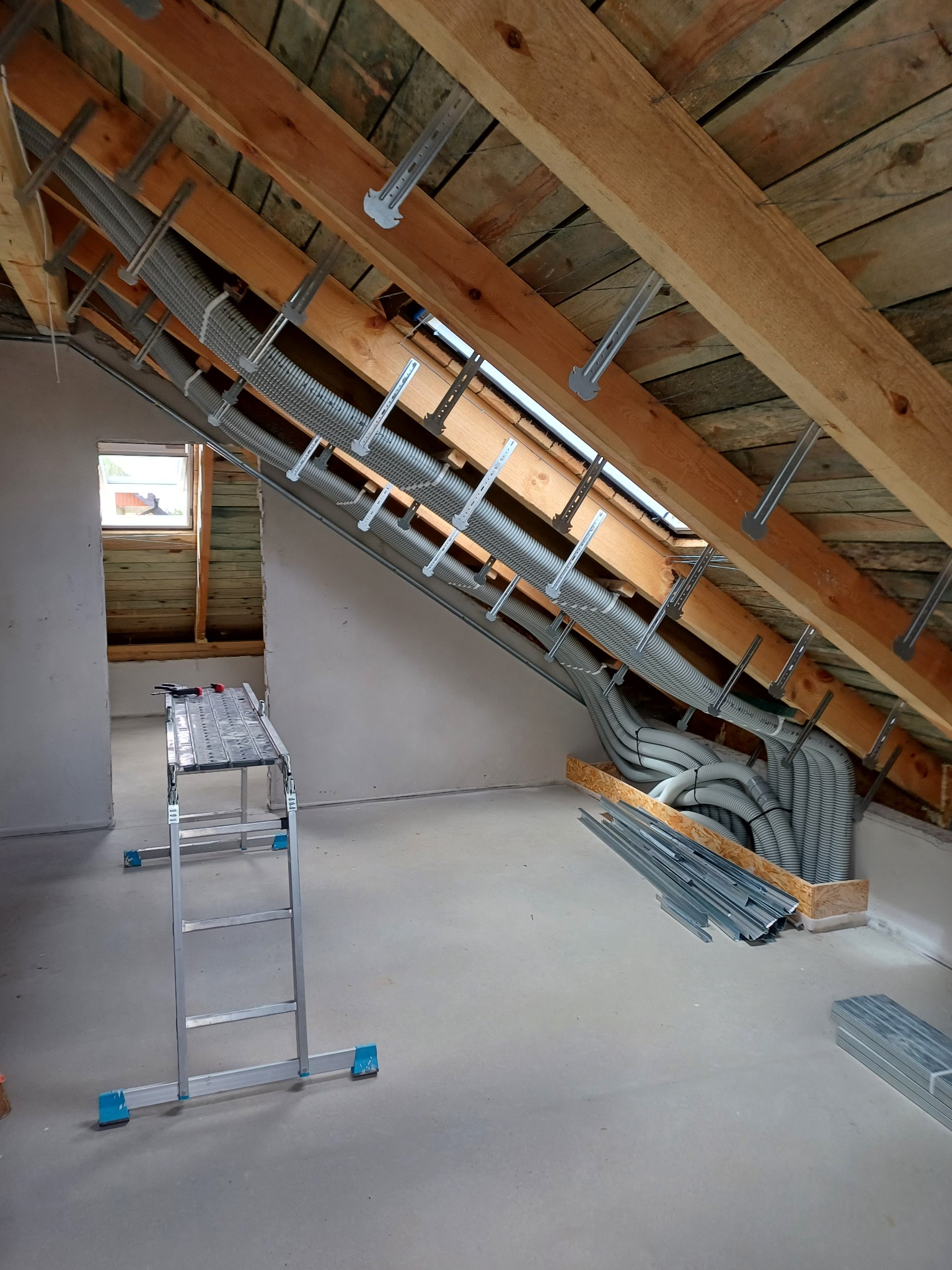 Poddasze w trakcie adaptacji: widoczne drewniane krokwie, metalowe profile mocujące i rury wentylacyjne. W tle okno i drabina. Surowy stan wykończenia.