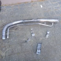 Metalowa rura paliwowa i uchwyty na szarym bruku. Elementy po demontażu, przygotowane do czyszczenia lub renowacji. Widoczne ślady użytkowania i korozji.