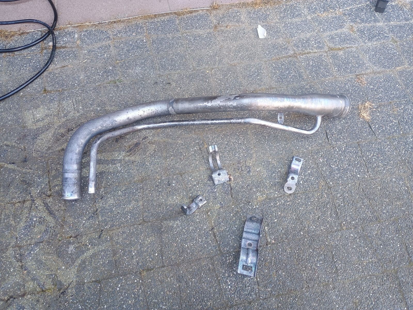 Metalowa rura paliwowa i uchwyty na szarym bruku. Elementy po demontażu, przygotowane do czyszczenia lub renowacji. Widoczne ślady użytkowania i korozji.