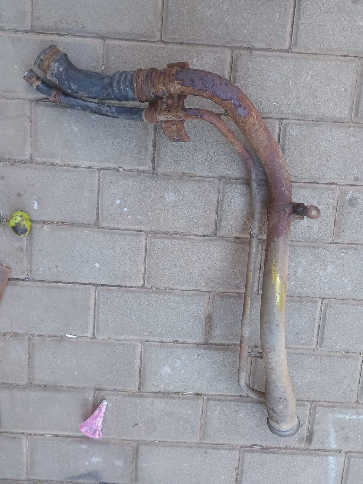 Zardzewiały, zakrzywiony element metalowy z gumowymi przewodami na brukowanej powierzchni. Widoczne ślady korozji i zużycia. Na ziemi leżą śmieci.