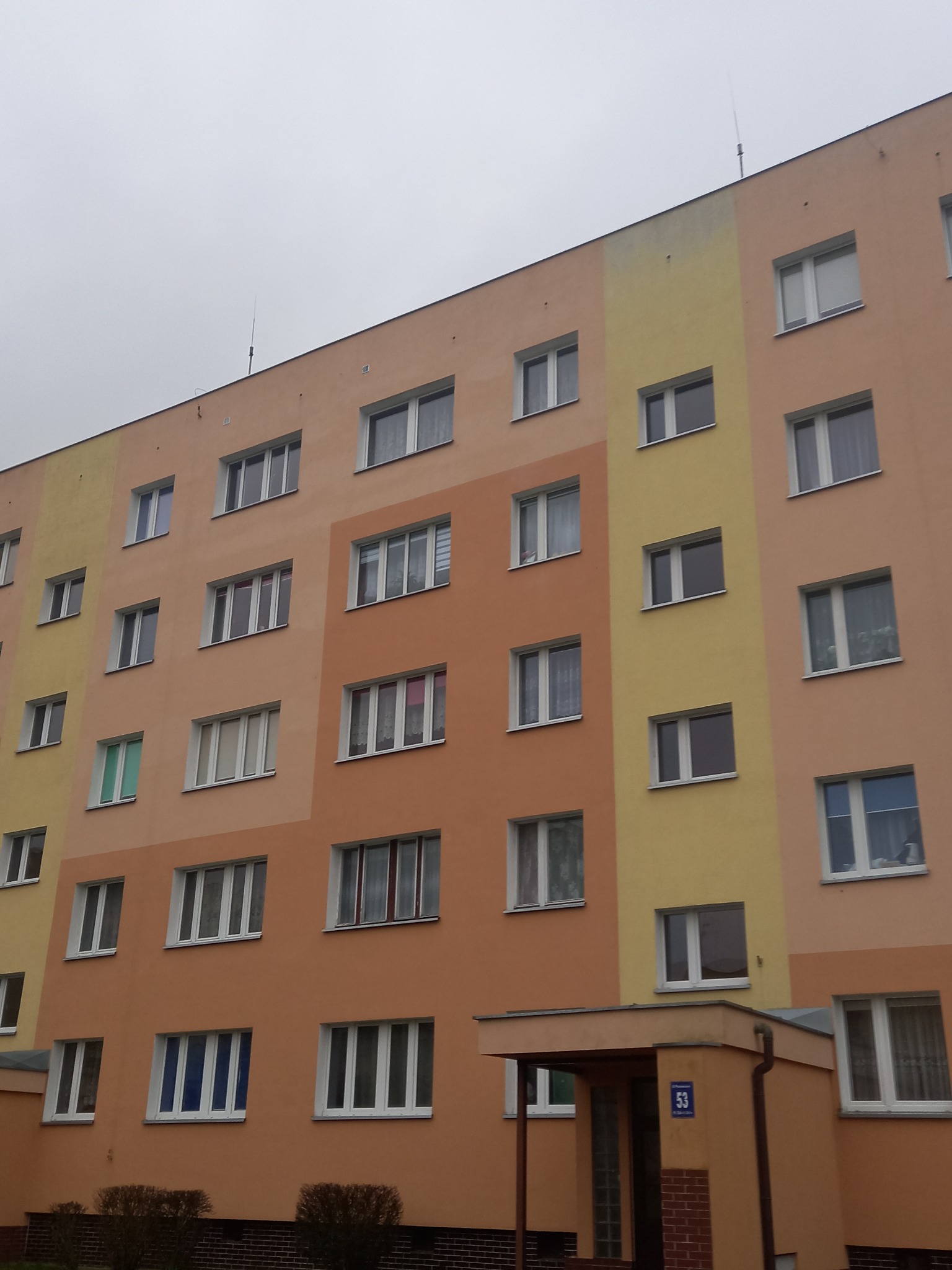 Wielopiętrowy budynek z numerem 53, pomalowany w odcieniach pomarańczy i żółci, z widocznymi oknami i wejściem. Pochmurne niebo w tle.