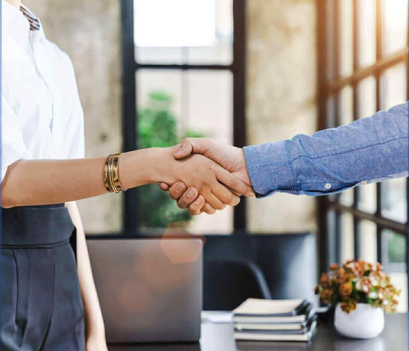 Uścisk dłoni kobiety z bransoletką i mężczyzny w błękitnej koszuli na tle biura z laptopem, symbolizujący partnerstwo i zaufanie w biznesie.