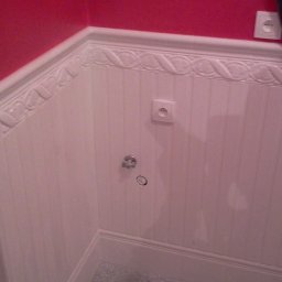 USŁUGI BUDOWLANE DARIUSZ RADOMSKI - Biała boazeria z dekoracyjnym frezem na ścianie łazienki, czerwona ściana u góry, gniazdka elektryczne i instalacja wodna. Widoczne odbicie osoby w lustrze.