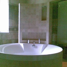 USŁUGI BUDOWLANE DARIUSZ RADOMSKI - Nowoczesna, okrągła wanna wolnostojąca w łazience wykończonej jasnymi płytkami. Widoczne okno i fragment kabiny prysznicowej. Minimalistyczny design.
