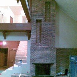 USŁUGI BUDOWLANE DARIUSZ RADOMSKI - Murowany kominek z cegły w surowym wnętrzu z drewnianą konstrukcją stropu i schodami. Widoczne narzędzia i materiały budowlane, etap prac wykończeniowych.