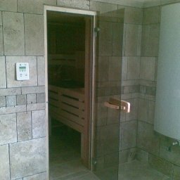USŁUGI BUDOWLANE DARIUSZ RADOMSKI - Wykończona sauna z kamienną okładziną ścian, szklanymi drzwiami i panelem sterowania. Widoczne drewniane ławy wewnątrz sauny i grzejnik na ścianie.