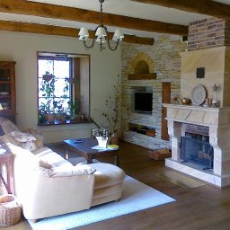 USŁUGI BUDOWLANE DARIUSZ RADOMSKI - Jasny salon z kamiennym kominkiem i drewnianymi belkami stropowymi. Okno z roślinami, drewniane meble i podłoga tworzą przytulne wnętrze.