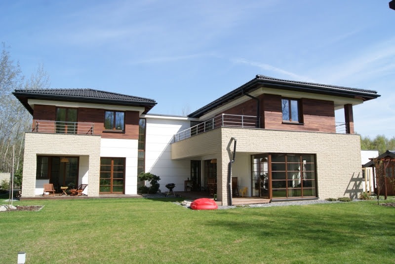 Dwukondygnacyjny dom z jasną elewacją i drewnianymi panelami, balkon z metalową balustradą, widok z zadbanego trawnika w słoneczny dzień.