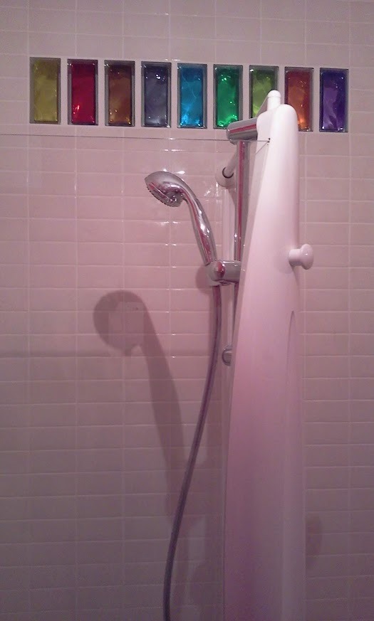 Biała kabina prysznicowa na tle ściany z jasnych płytek, urozmaiconej poziomym pasem kolorowych, szklanych elementów dekoracyjnych. Widoczna słuchawka prysznicowa.