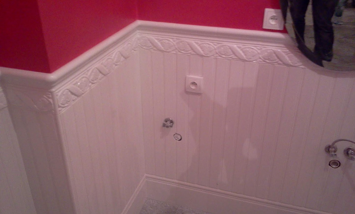 Biała boazeria z dekoracyjnym frezem na ścianie łazienki, czerwona ściana u góry, gniazdka elektryczne i instalacja wodna. Widoczne odbicie osoby w lustrze.