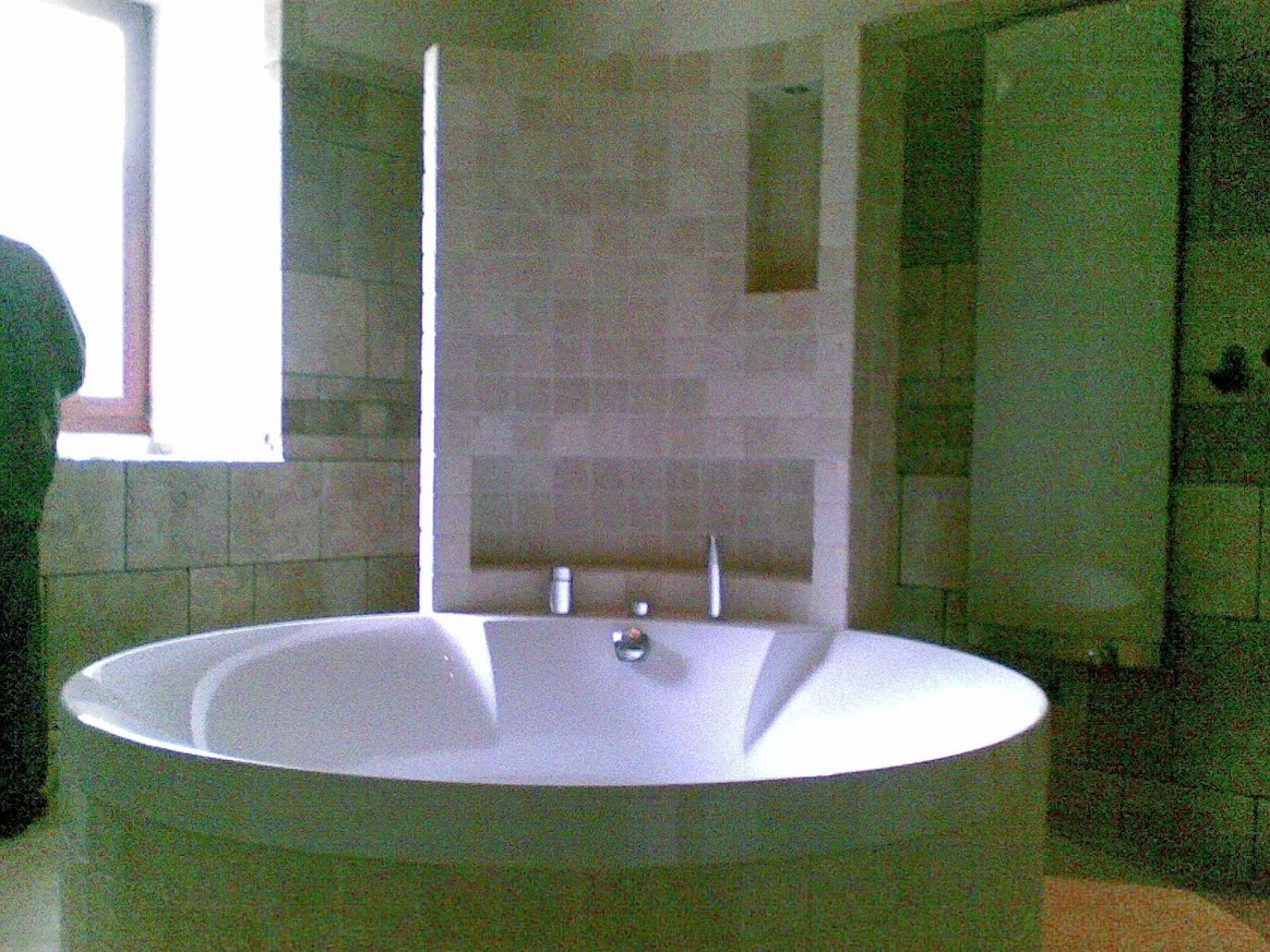 Nowoczesna, okrągła wanna wolnostojąca w łazience wykończonej jasnymi płytkami. Widoczne okno i fragment kabiny prysznicowej. Minimalistyczny design.