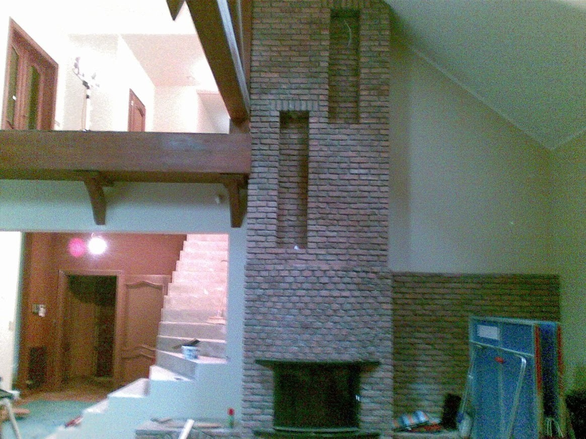 Murowany kominek z cegły w surowym wnętrzu z drewnianą konstrukcją stropu i schodami. Widoczne narzędzia i materiały budowlane, etap prac wykończeniowych.