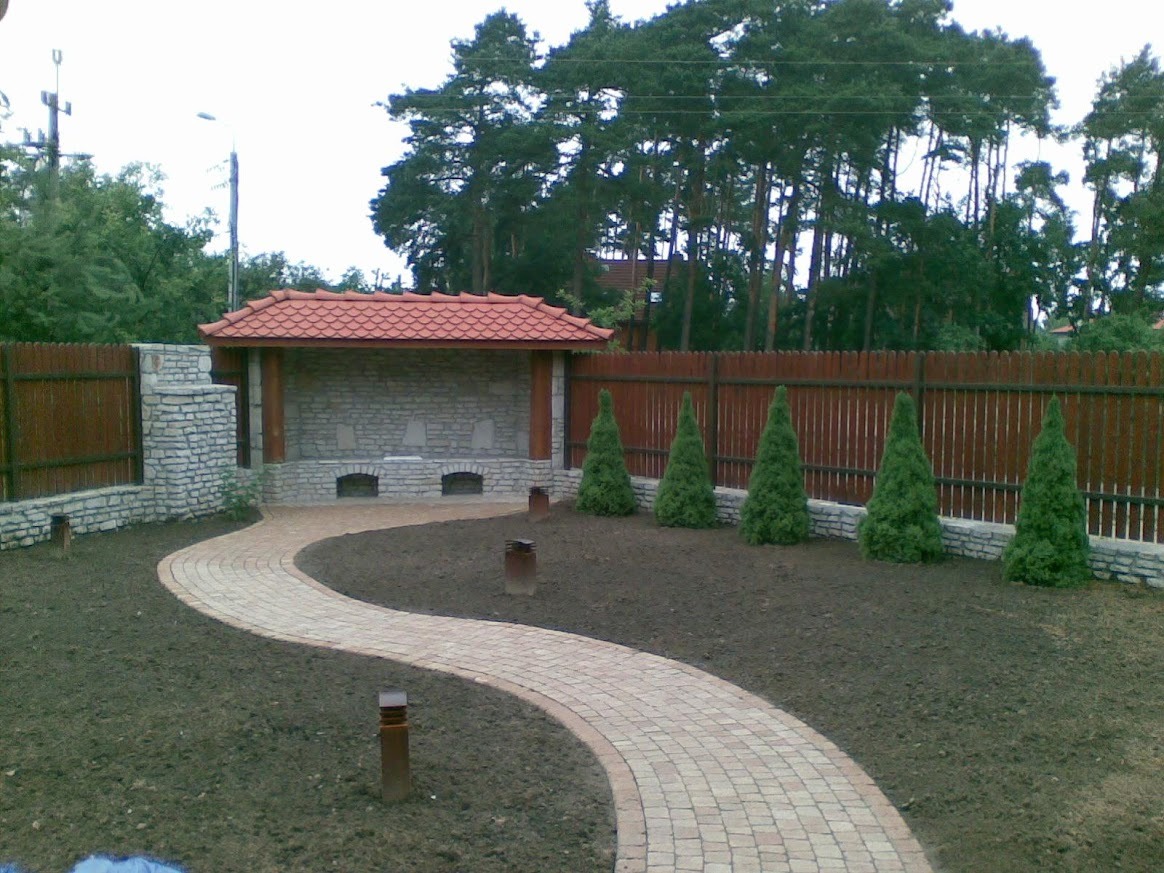 Kamienna altana z czerwonym dachem, brukowana ścieżka i drewniany płot w ogrodzie. Widok na drzewa w tle. Elementy małej architektury ogrodowej.