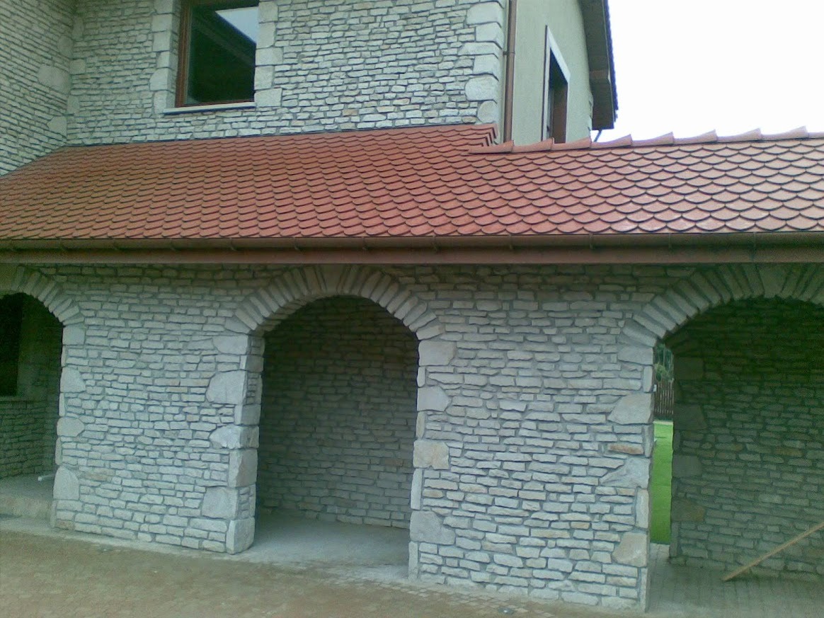 Kamienna elewacja budynku z trzema łukowymi przejściami i czerwoną dachówką. Widoczna faktura kamienia i detale architektoniczne.