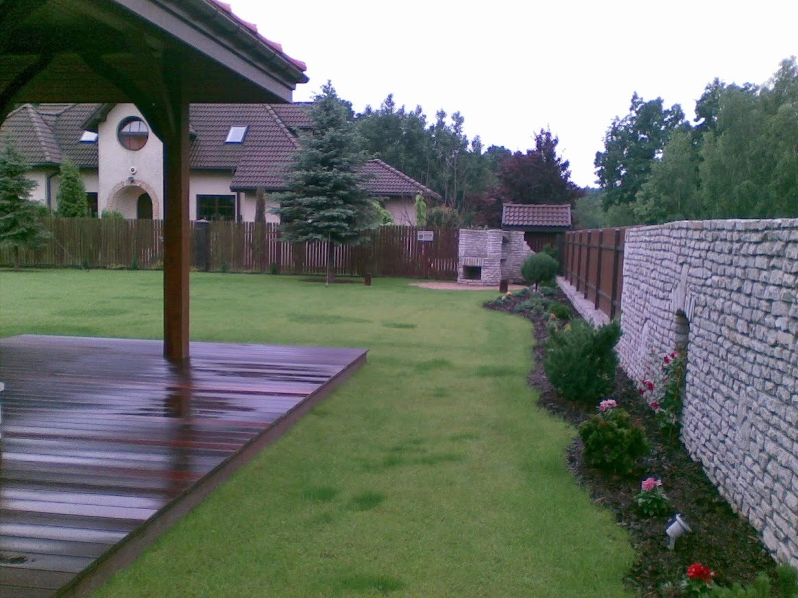 Widok na zadbany ogród z drewnianym tarasem, kamiennym murem i grillem. W tle dom z brązowym dachem i ogrodzeniem. Całość w spokojnej, zielonej tonacji.