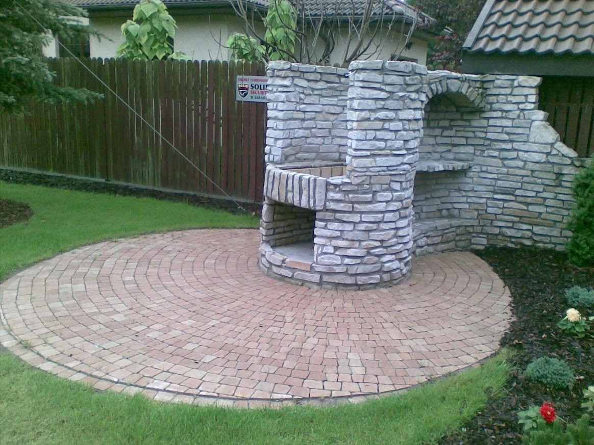Murowany grill z kamienia na okrągłym patio z kostki, otoczony trawnikiem i roślinami. W tle drewniany płot i budynek z dachówką.