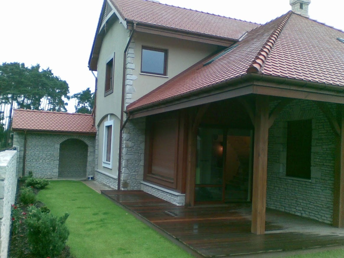 Elewacja domu z kamienną okładziną, drewnianym tarasem i zadaszeniem. Czerwona dachówka i zadbany trawnik dopełniają całość.