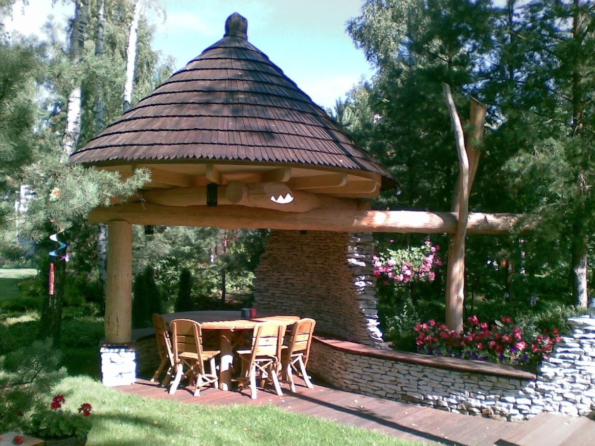 Drewniana altana ogrodowa z kamiennymi elementami, kryta gontem, z okrągłym stołem i krzesłami wewnątrz, otoczona zielenią i kwiatami.