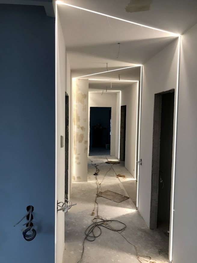 Korytarz w trakcie prac wykończeniowych z zamontowanym oświetleniem LED wzdłuż ścian i sufitu. Widoczne przewody i niedokończone ściany.
