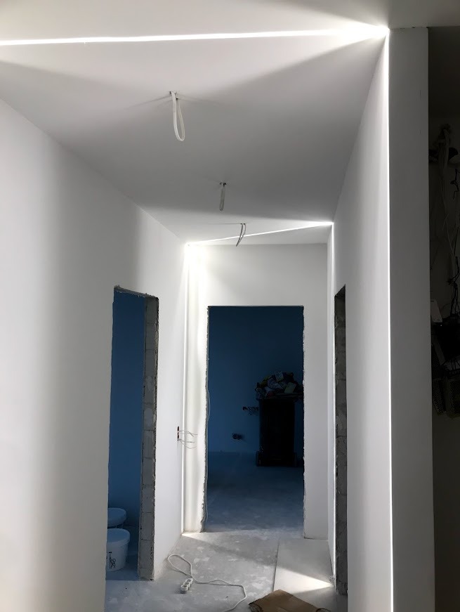 Białe wnętrze w trakcie wykańczania z oświetleniem LED wzdłuż sufitu i otworów drzwiowych. Widoczne niedoskonałości i surowy stan podłogi.