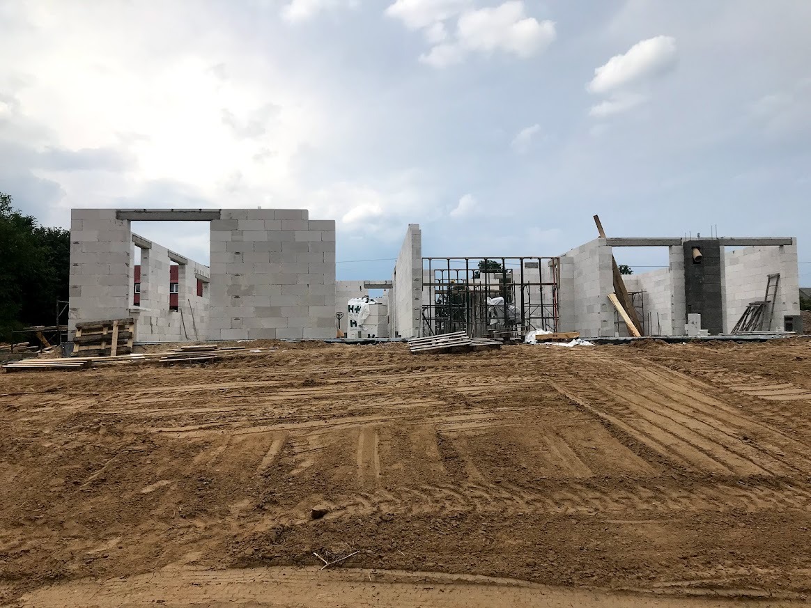 Surowy stan budynku w trakcie budowy z białych bloczków, widoczne otwory okienne i drzwiowe, teren budowy z ziemią i śladami opon, pochmurne niebo.