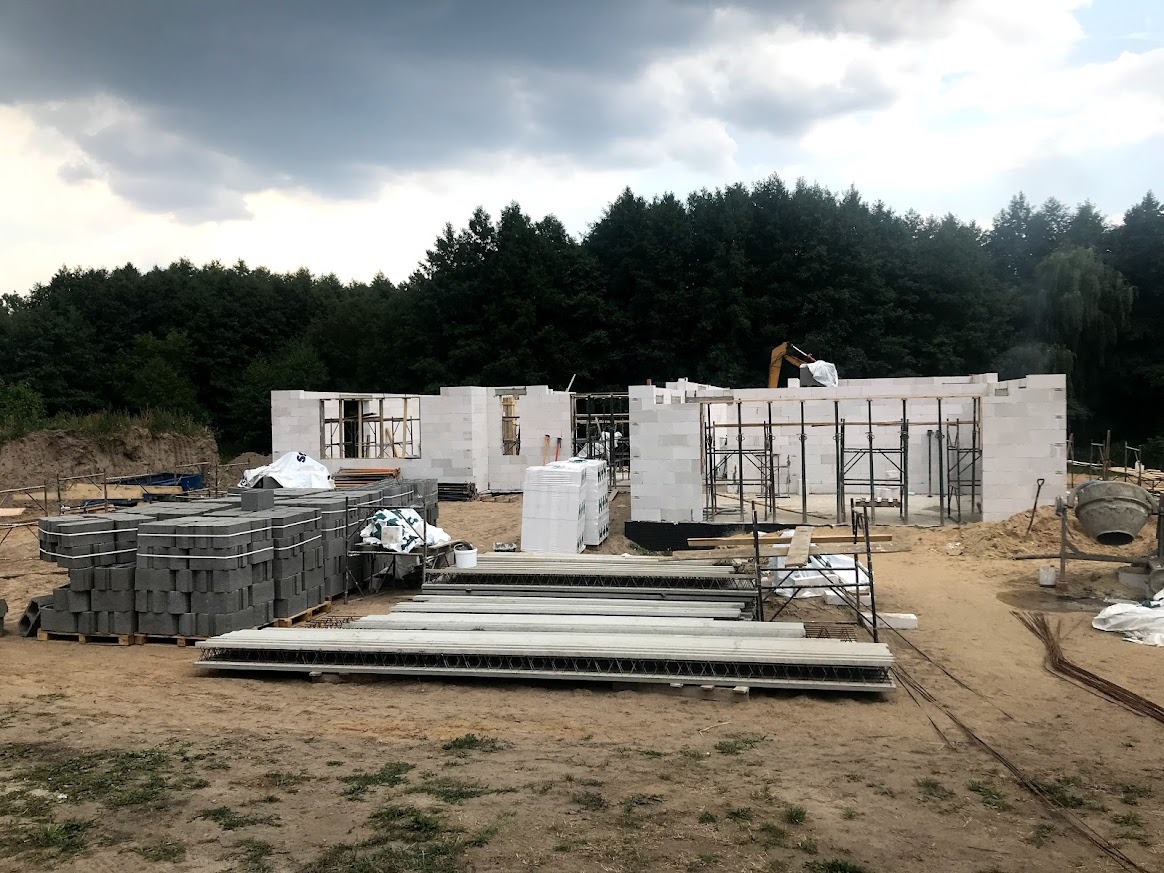 Budowa domu w trakcie wznoszenia ścian z bloczków betonu komórkowego. Widoczne rusztowania, materiały budowlane i częściowo zarys lasu w tle.