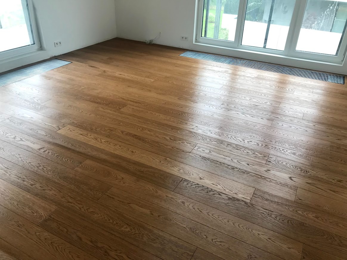 Ukończona podłoga z paneli drewnianych w jasnym pomieszczeniu z dużymi oknami. Widoczne grzejniki pod oknami. Naturalne oświetlenie podkreśla strukturę drewna.