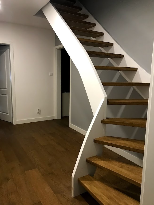 Nowoczesne, drewniane schody z białymi elementami konstrukcyjnymi, prowadzące na wyższą kondygnację. Widoczna podłoga z paneli w odcieniach brązu i białe ściany.