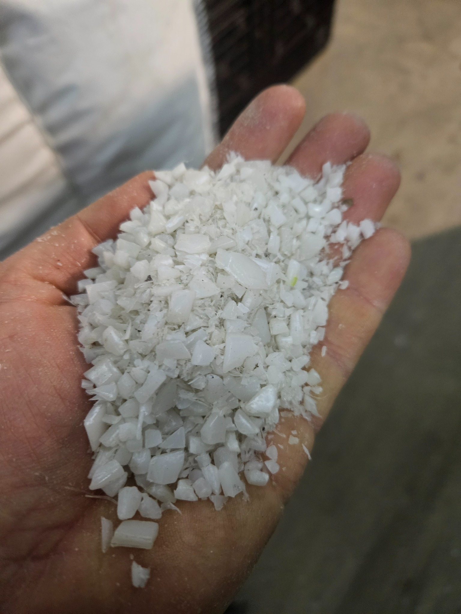 Dłoń trzymająca biały granulat, prawdopodobnie tworzywo sztuczne. Widoczny worek w tle. Materiał o nieregularnych kształtach i matowej powierzchni.