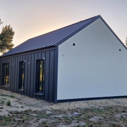 DervoBud Andrii Holdyshchuk - Nowoczesny dom z ciemną elewacją z blachy trapezowej i jasną, gładką ścianą szczytową. Widoczne okna i metalowy dach na tle drzew i zachodzącego słońca.