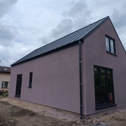 DervoBud Andrii Holdyshchuk - Nowoczesny, jednopiętrowy dom z ciemną blachą na dachu i elewacją w odcieniach fioletu. Widoczne czarne ramy okien i rynna. Na pierwszym planie ziemia.