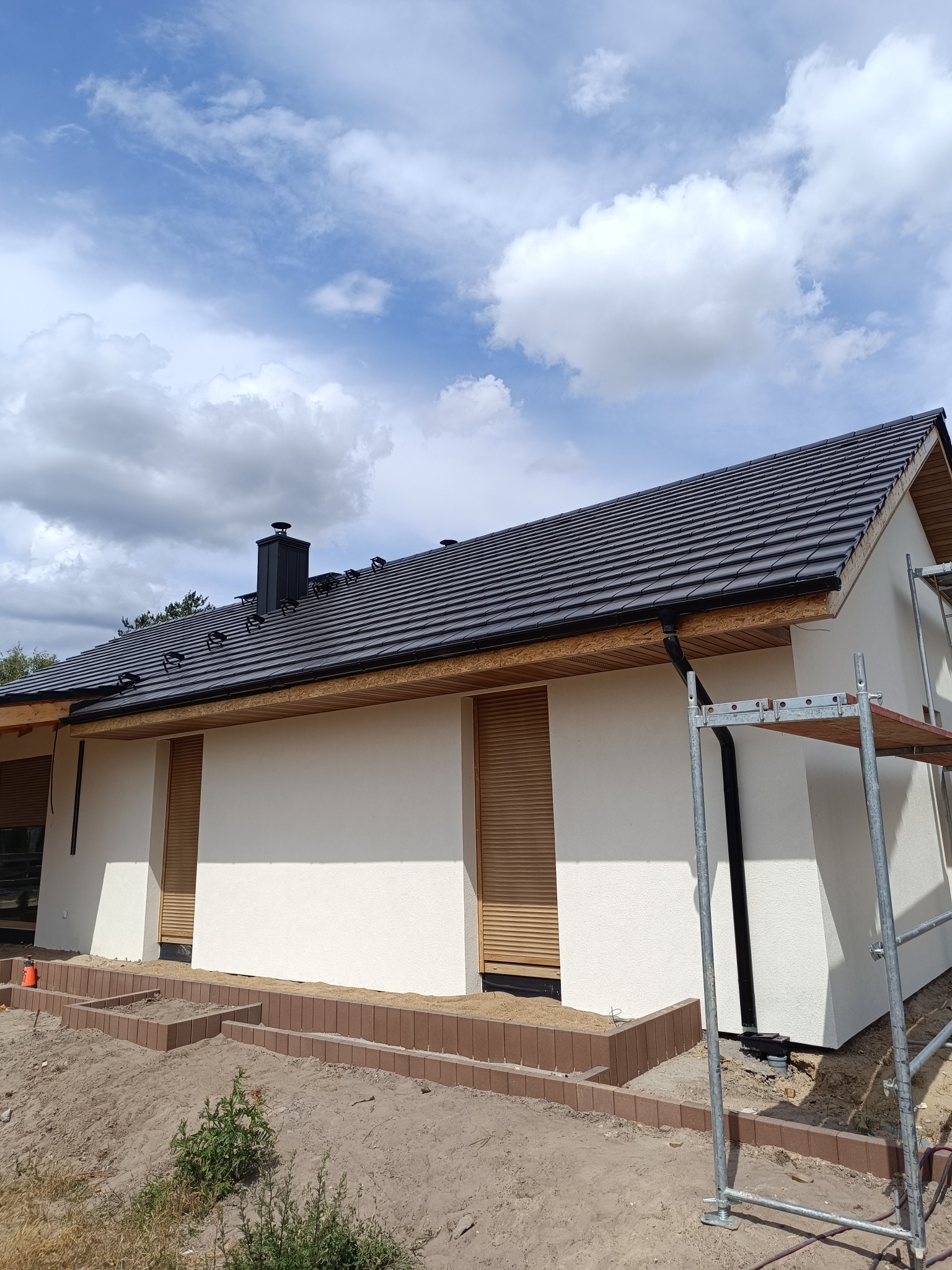 Niski, biały dom z ciemnym dachem i drewnianymi elementami wykończeniowymi. Widoczne rusztowanie i częściowo ułożona kostka brukowa wokół budynku.