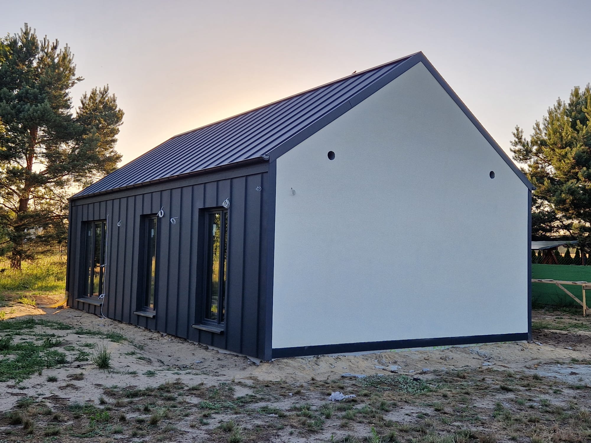 Nowoczesny dom z ciemną elewacją z blachy trapezowej i jasną, gładką ścianą szczytową. Widoczne okna i metalowy dach na tle drzew i zachodzącego słońca.