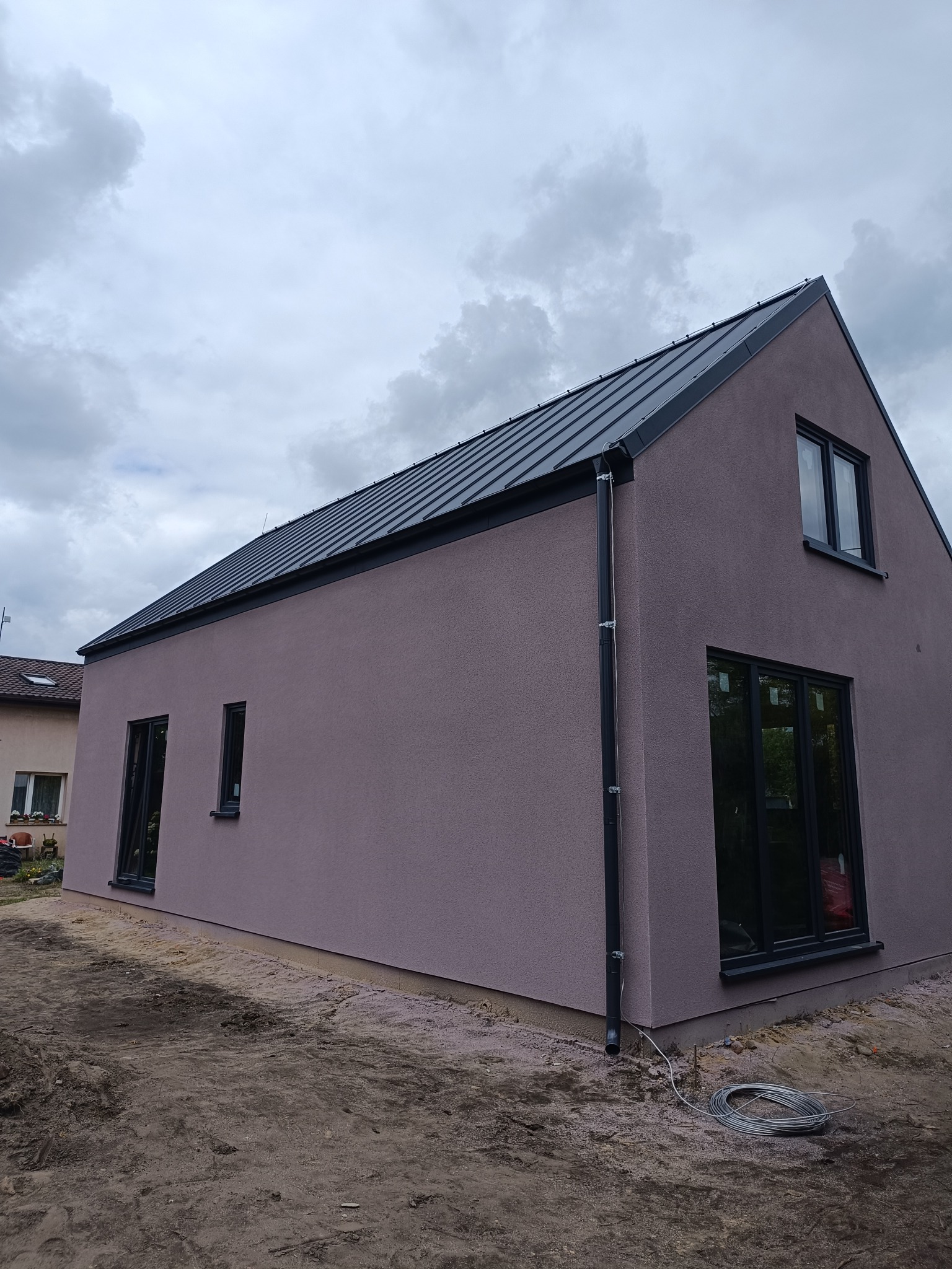 Nowoczesny, jednopiętrowy dom z ciemną blachą na dachu i elewacją w odcieniach fioletu. Widoczne czarne ramy okien i rynna. Na pierwszym planie ziemia.