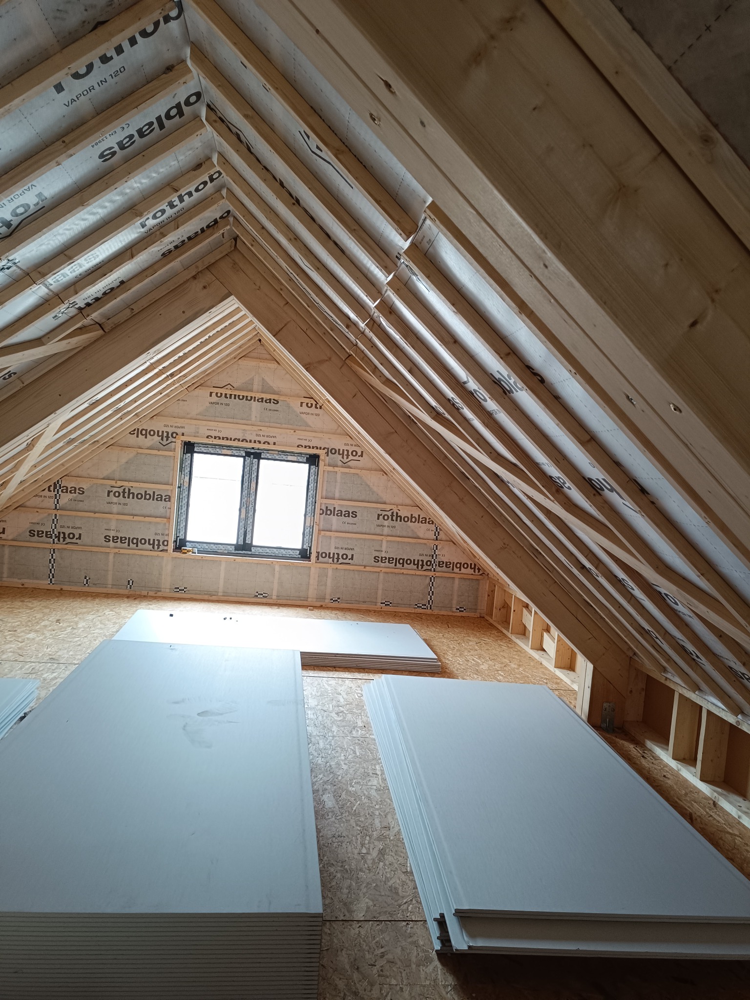 Wnętrze poddasza z drewnianą konstrukcją szkieletową, oknem i przygotowanymi płytami. Widoczne zabezpieczenie folią paroizolacyjną.