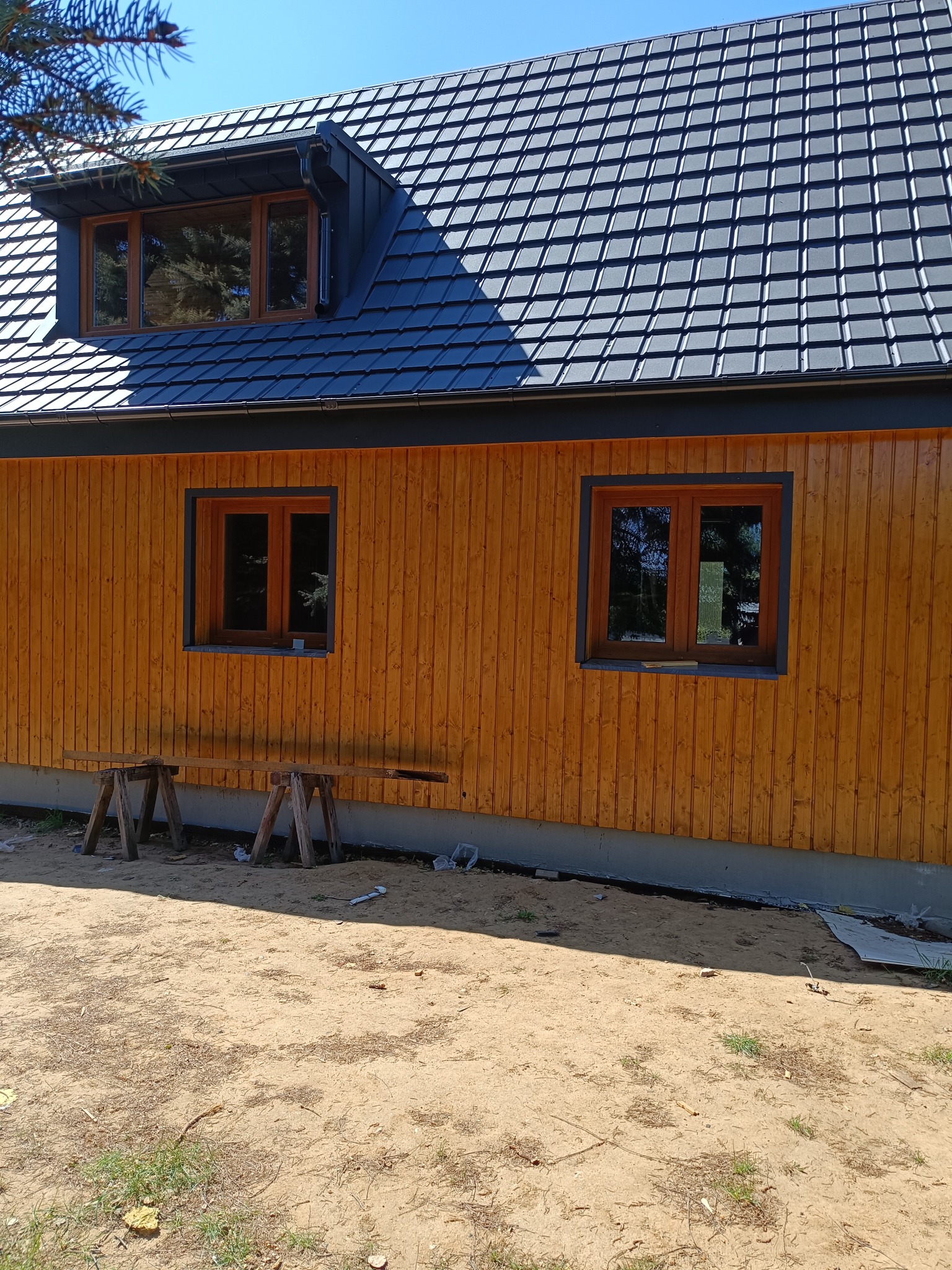 Drewniana elewacja nowo wybudowanego domu z ciemną dachówką i drewnianą stolarką okienną. Na pierwszym planie dwa drewniane kozły.