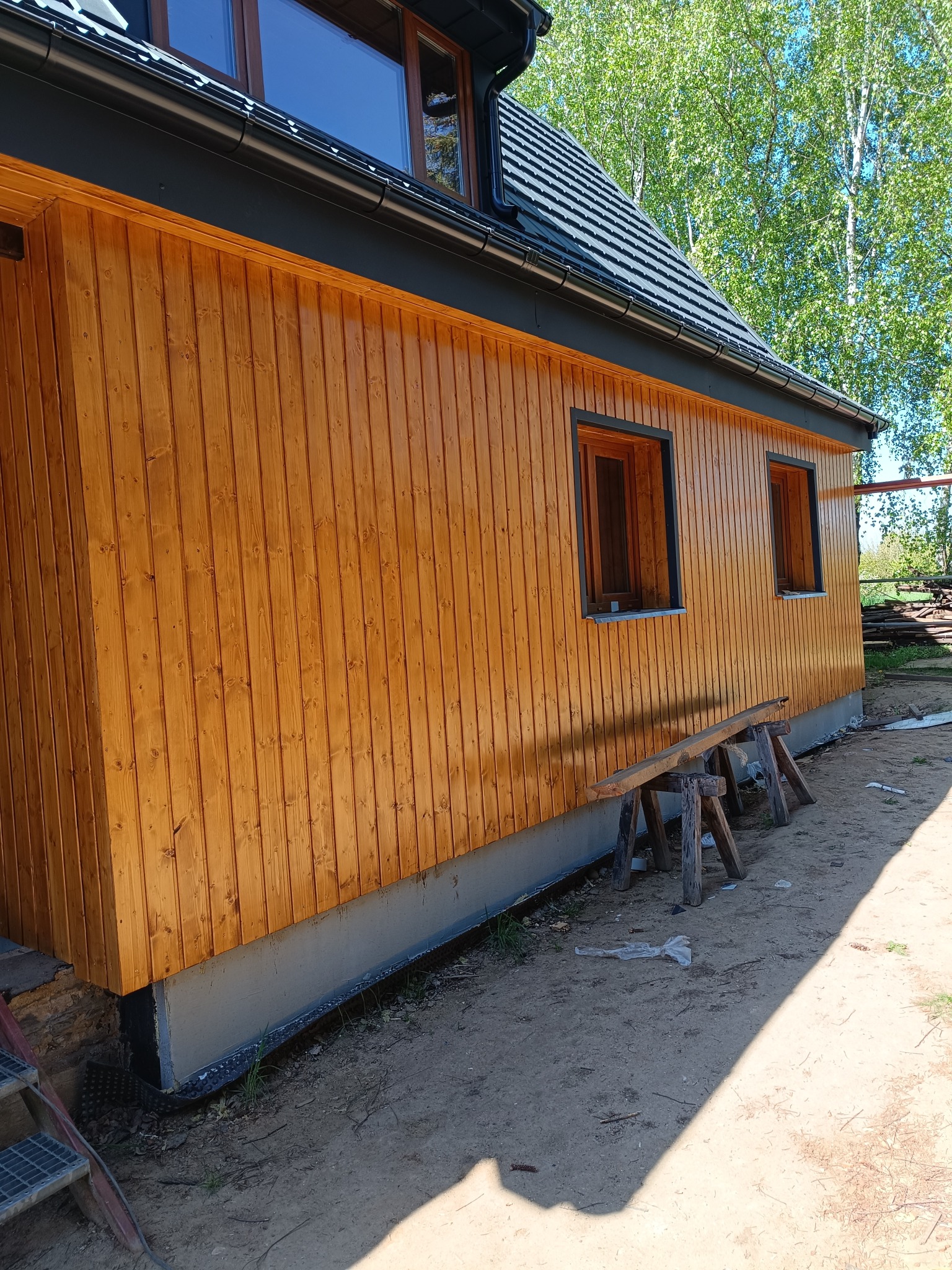 Nowoczesny dom z drewnianą elewacją w odcieniach brązu. Widoczne okna z czarnymi ramami, ciemna dachówka i fragment zieleni w tle. Ujęcie z boku.