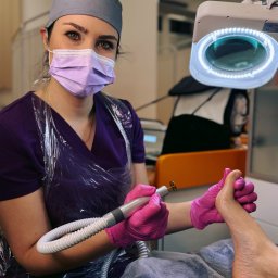 FOOT CARE PIONEERS Olga Legkoszkur - Podolog w fioletowym uniformie, w masce i rękawiczkach, opracowuje paznokcie frezarką pod lampą powiększającą. Zabieg pedicure w gabinecie kosmetycznym.