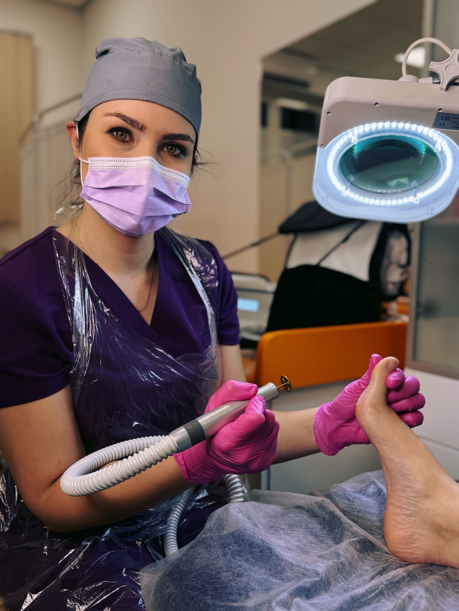Podolog w fioletowym uniformie, w masce i rękawiczkach, opracowuje paznokcie frezarką pod lampą powiększającą. Zabieg pedicure w gabinecie kosmetycznym.