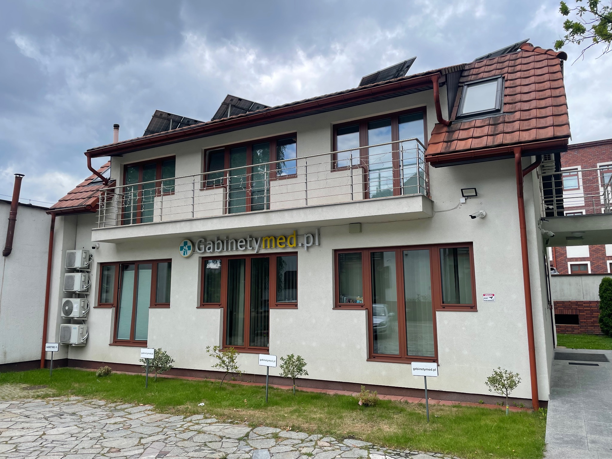 Dwukondygnacyjny budynek kliniki z balkonem i panelami słonecznymi na dachu, widoczny z zewnątrz, z tabliczkami informacyjnymi na trawniku przed budynkiem.