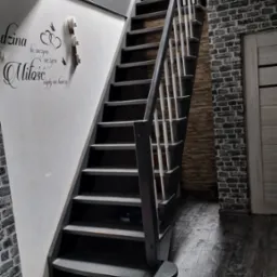 Schody w ciemnej tonacji z białymi balustradami, ściana z dekoracyjnym napisem, imitacja cegły na ścianach i podłodze, oświetlenie punktowe w suficie.