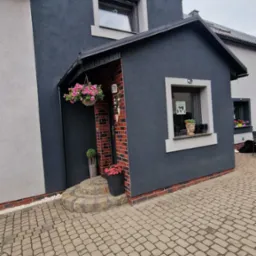 Elewacja domu z ciemnoszarą strukturą, ceglanym obramowaniem i jasnymi oknami. Wejście z doniczkami i wiszącymi kwiatami. Brukowany podjazd.